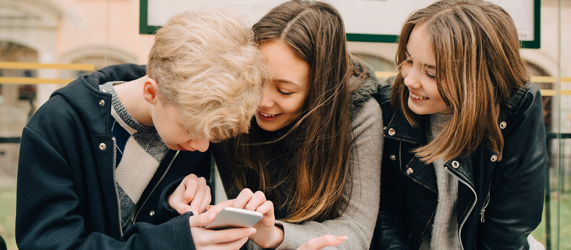 Tre tonåringar sitter på en busshållplats och tittar på en mobil