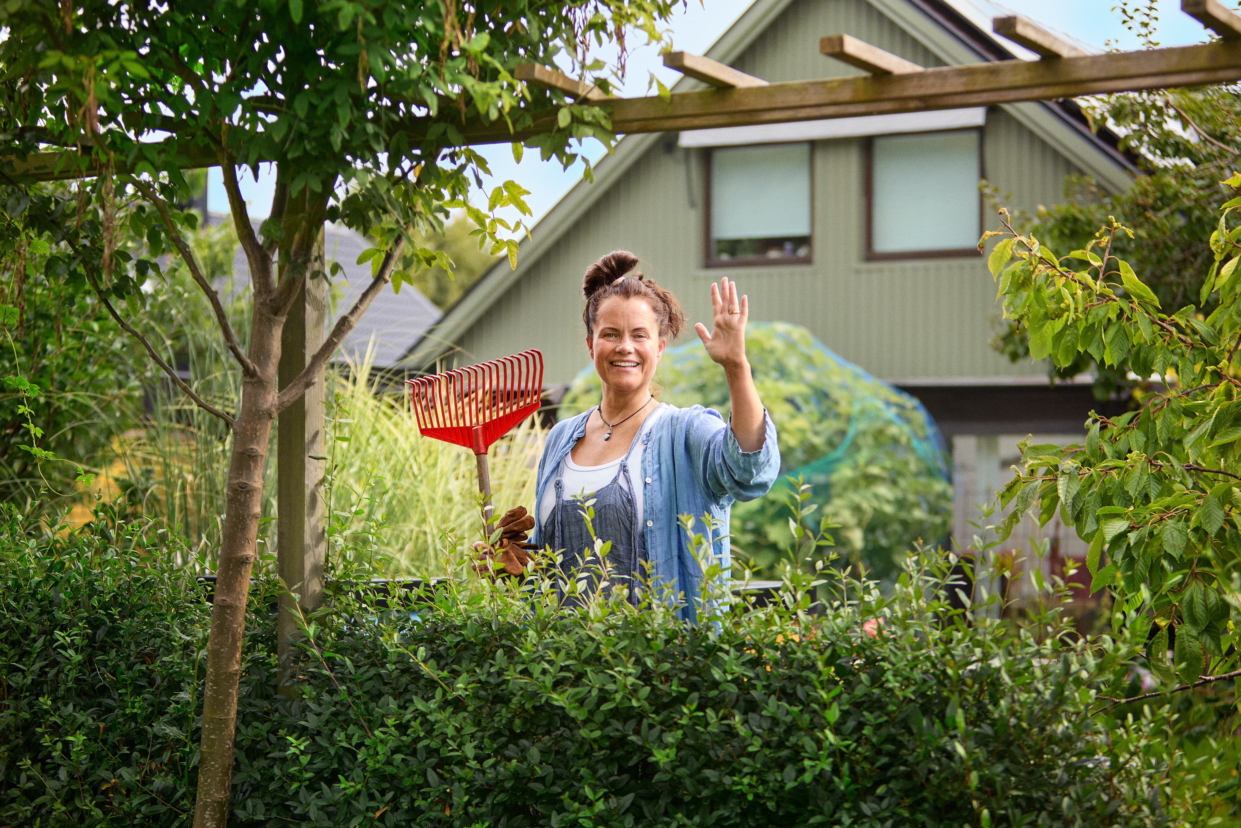 Kvinna med räfsa står bakom en häck. 